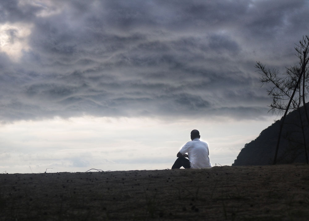 An image of loneliness. A man sits on a bare hill staring at a cloudy sky.