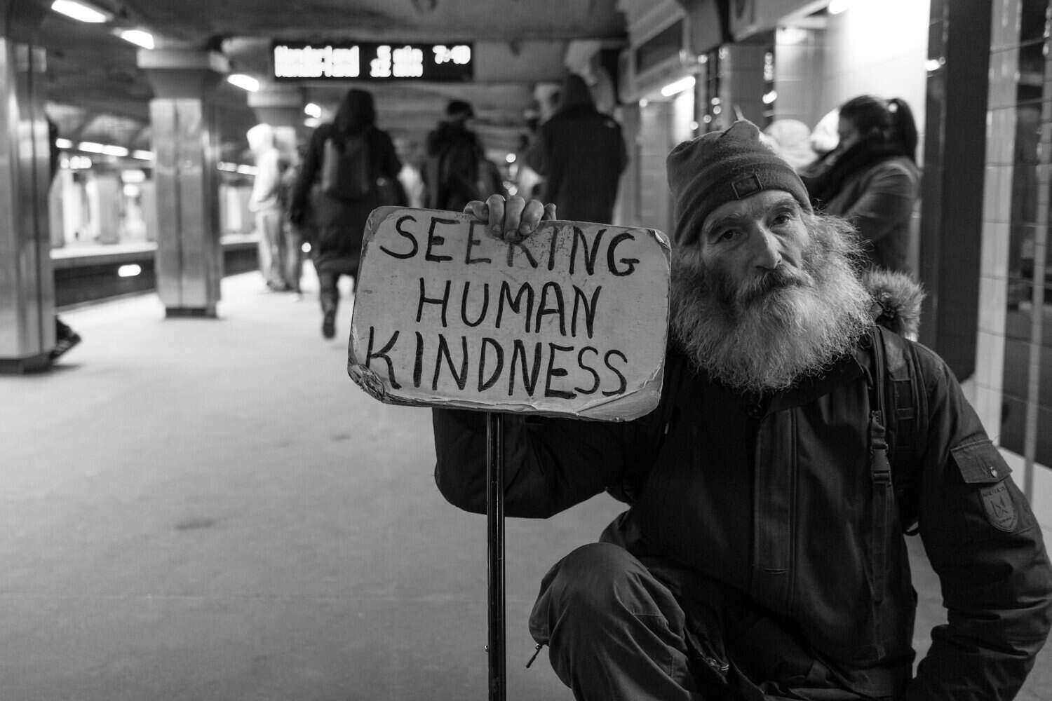A homeless man with a hand-made sign that asks for human kindness.